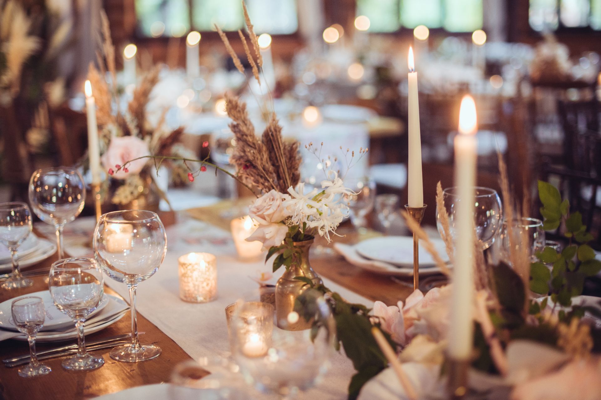 Elegantly set dining tables with lit candles, floral arrangements, and glassware in a warm, rustic-themed reception room.