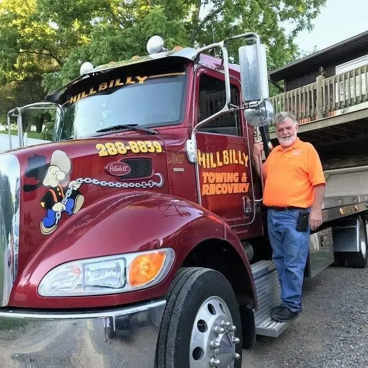 Maroon Hillbilly Towing & Recovery truck parked outdoors, with a man standing beside it.