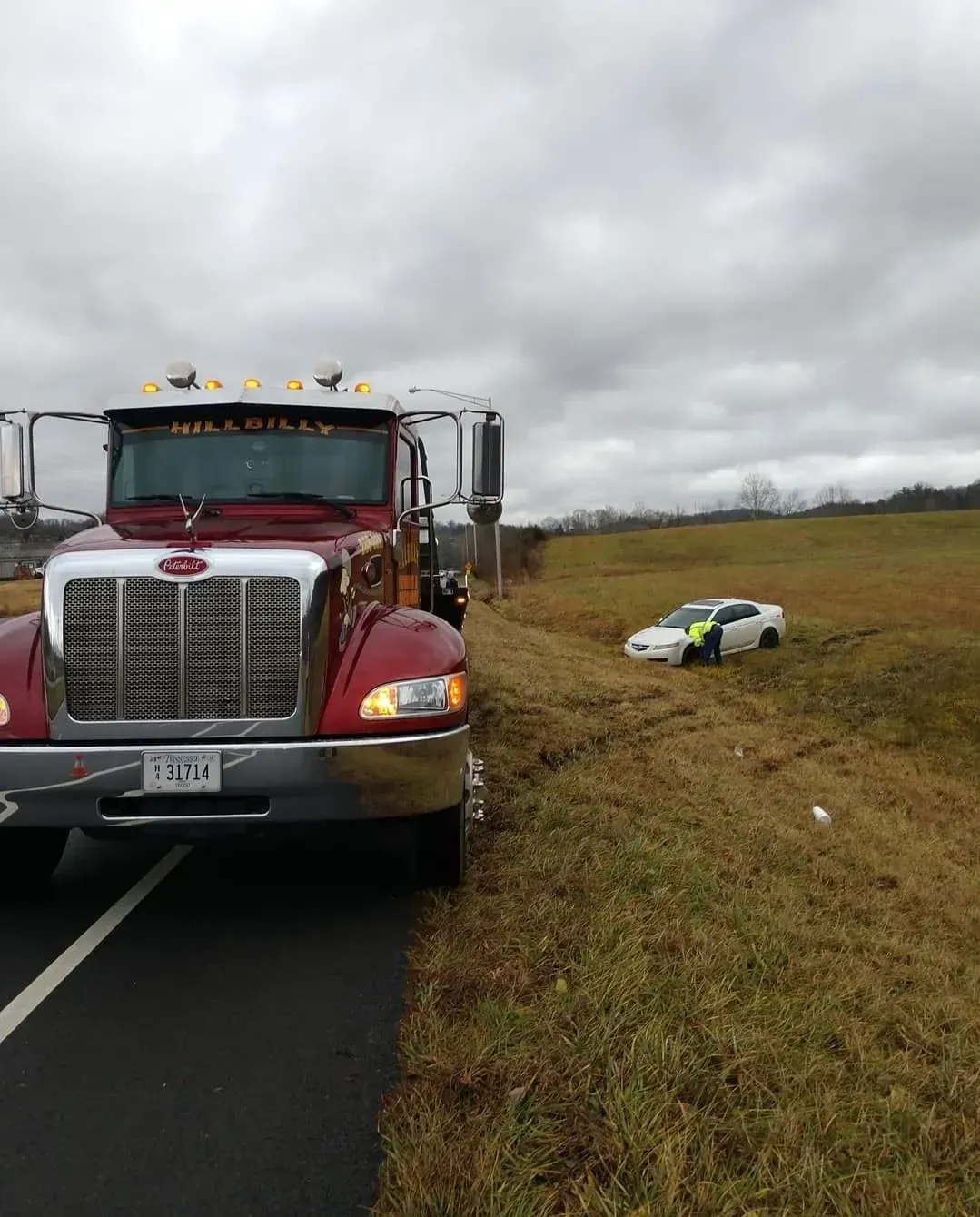 Semi-truck on road shoulder beside a small white car in a grassy ditch under cloudy skies.