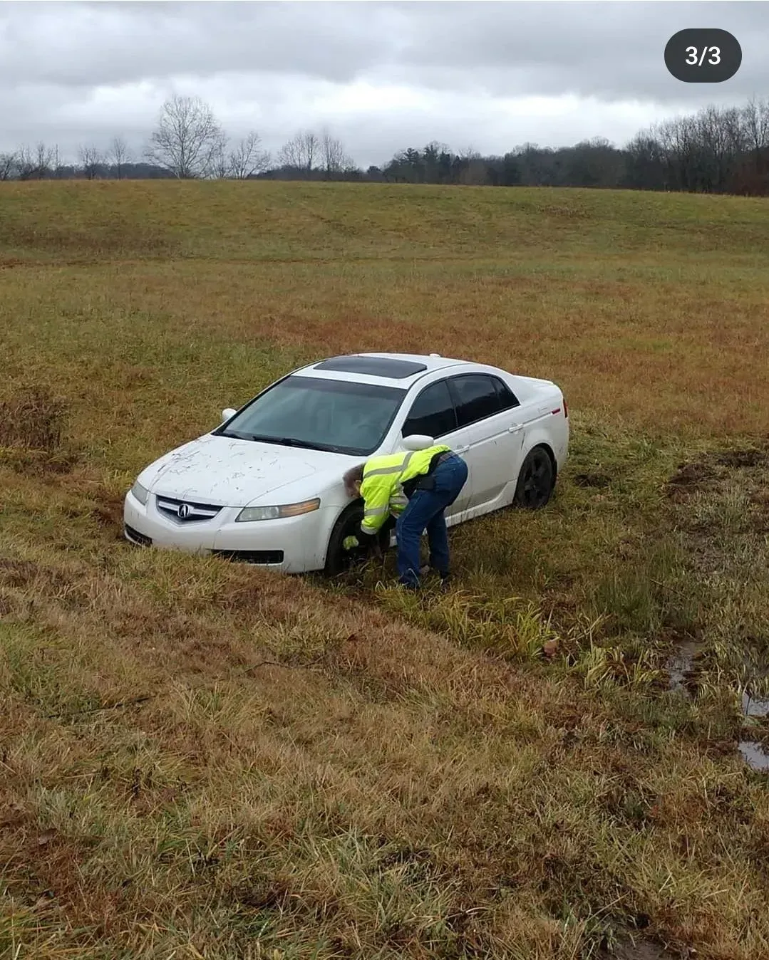 White car stuck in a muddy field, with a person in a bright jacket pushing from the front.