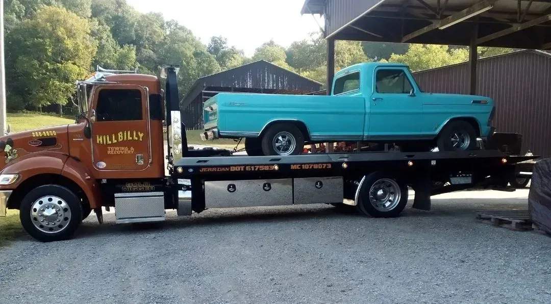 Tow truck hauling a turquoise vintage pickup truck in a driveway, with a dark building behind it.