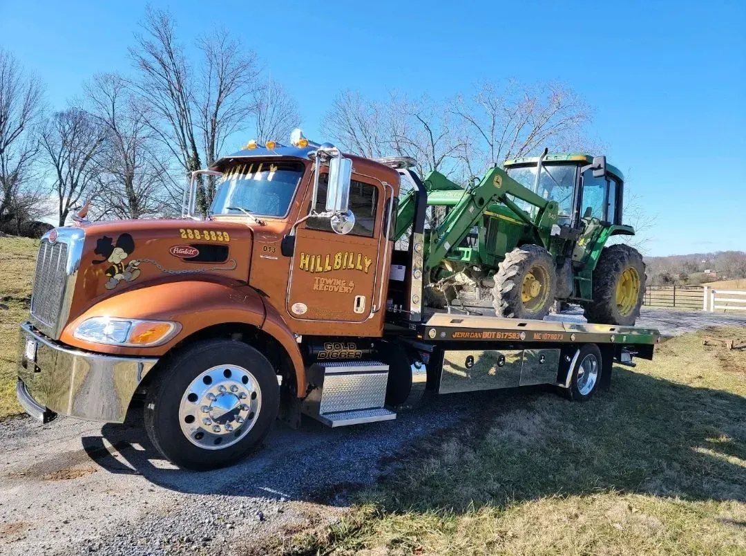 Orange tow truck hauling a green tractor in a grassy rural field under a clear blue sky