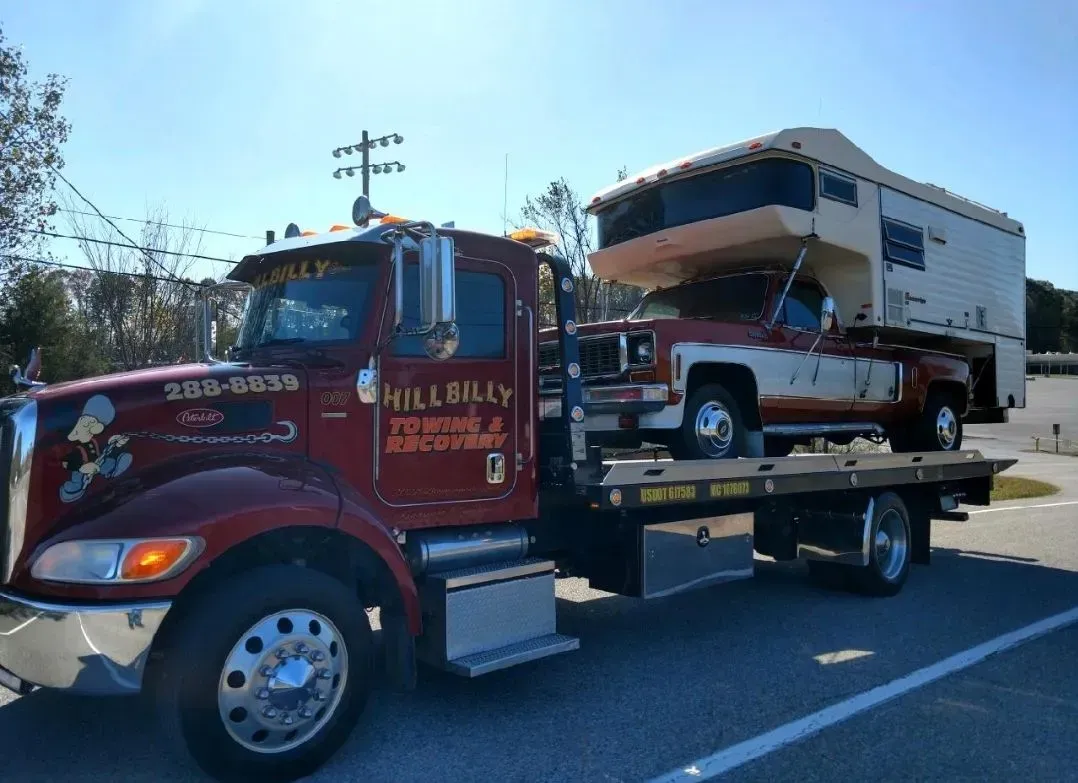 Red tow truck hauling a white pickup on a flatbed along a highway under a blue sky