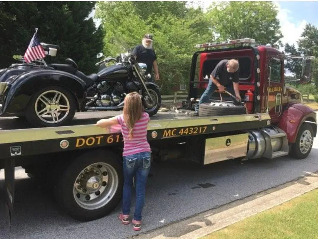 Tow truck carrying a black motorcycle and an off-road vehicle while a child watches on the roadside