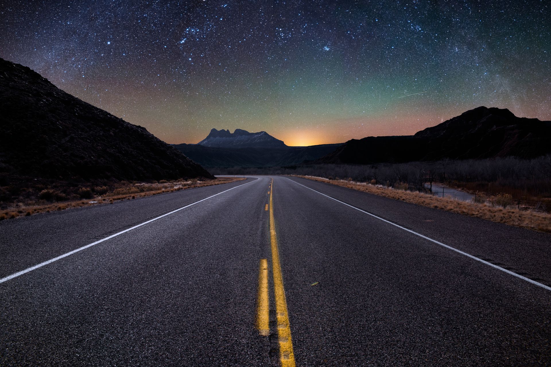 Empty road at night under a starry sky, with yellow center lines leading toward distant mountains.