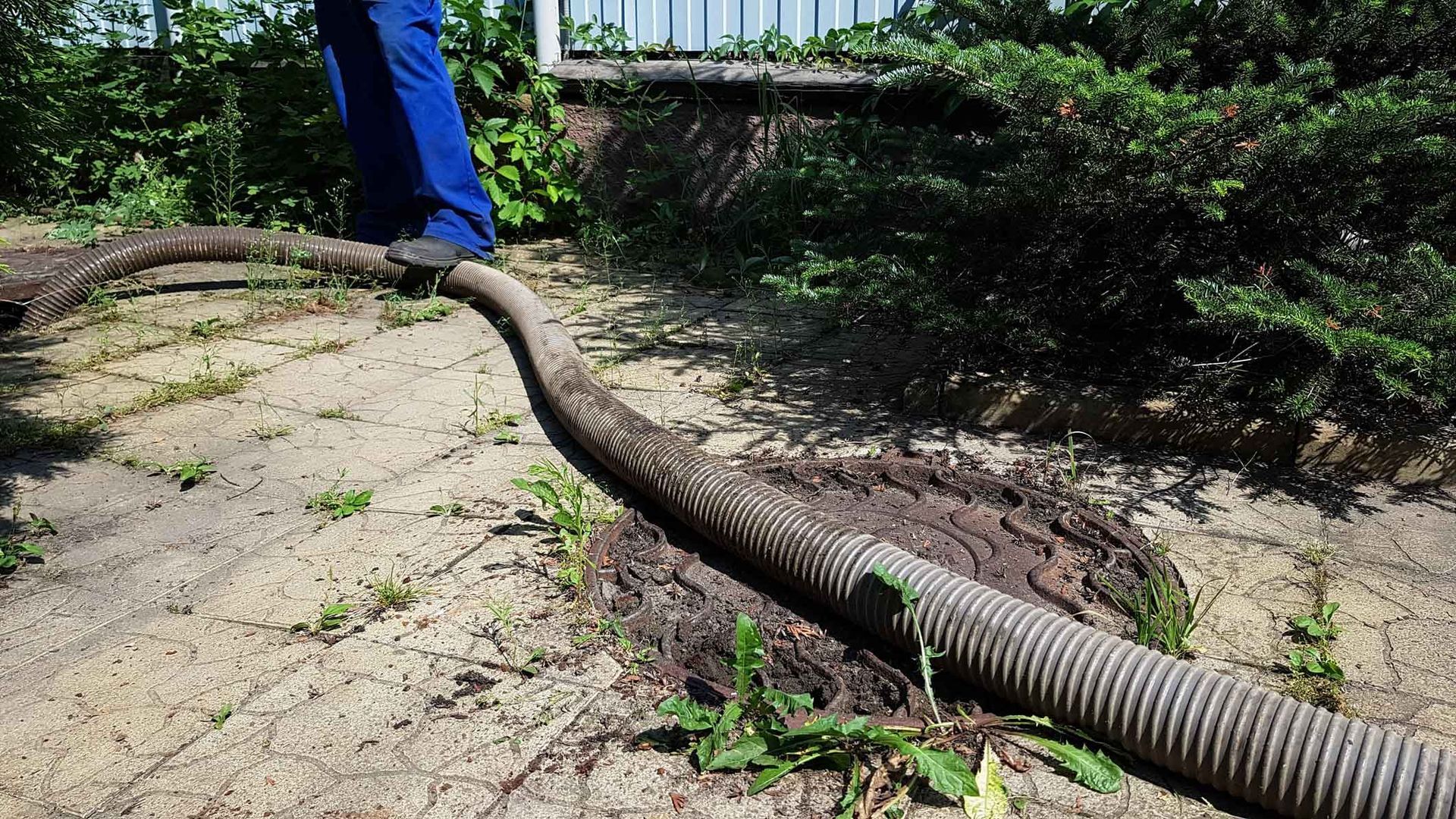 Ribbed vacuum hose draining an open septic tank manhole on a cracked concrete yard beside a worker.
