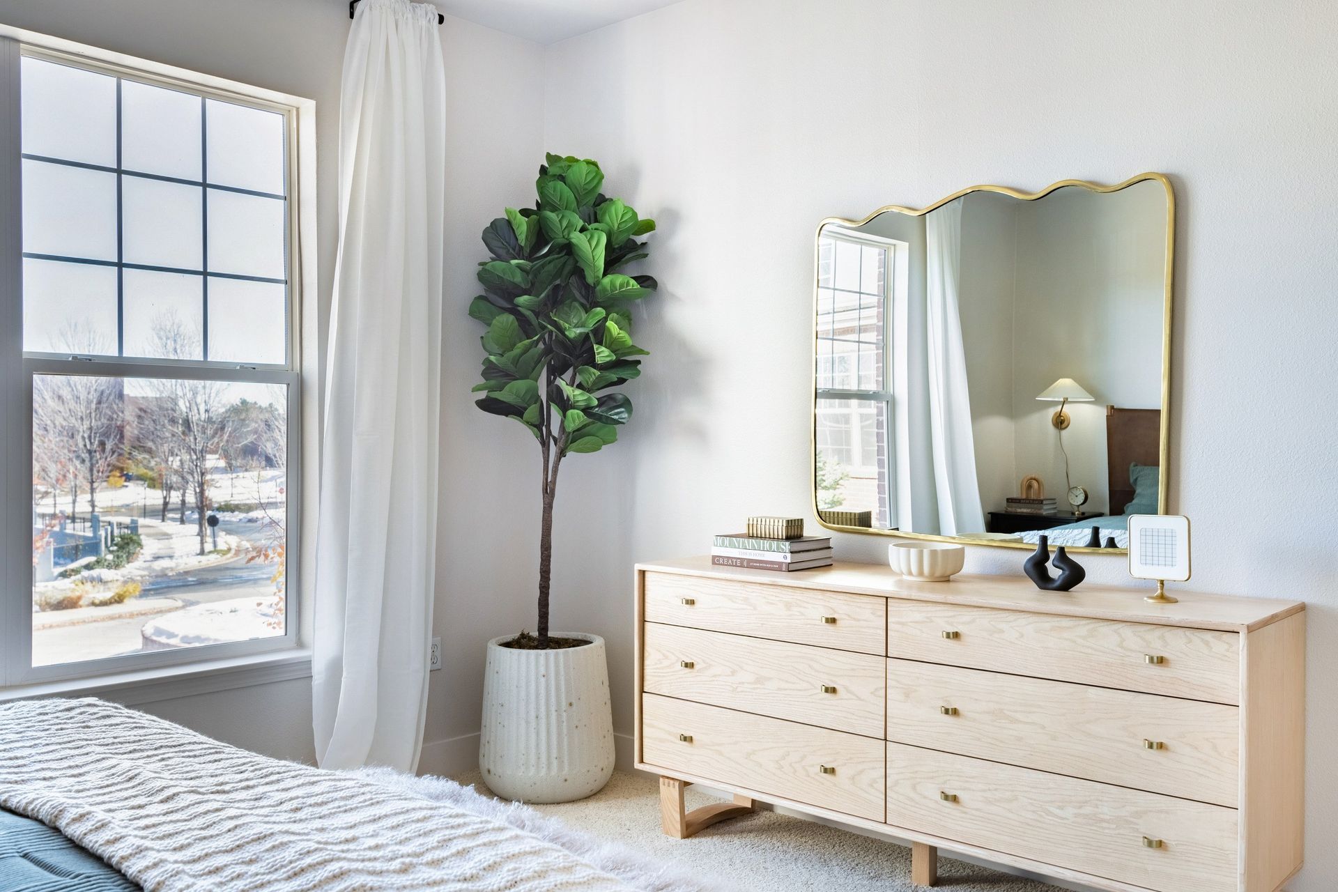 Bedroom with dresser, mirror, and fiddle-leaf fig plant. Light wood, white walls, and window with a view at The Viridian in Greenwood Village, CO.