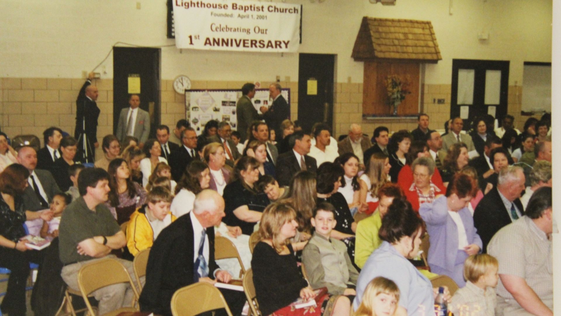 Crowded indoor anniversary event with seated guests applauding under a banner and brick walls.
