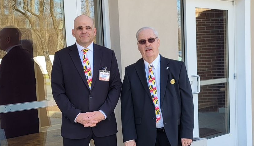 Two men in dark suits stand outside a building entrance, one wearing sunglasses.