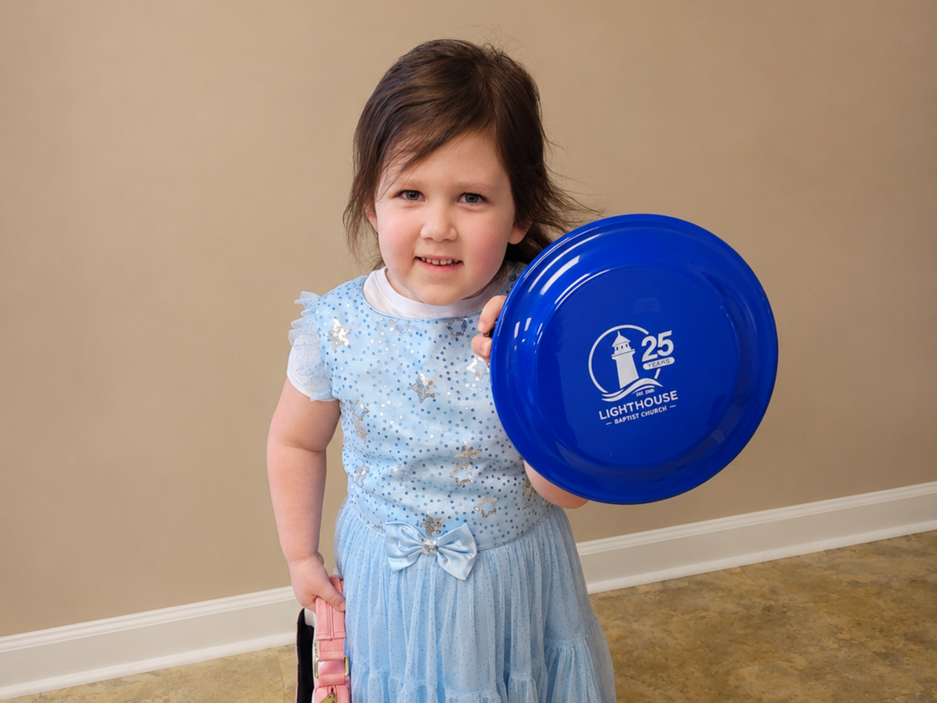 Young child in a light blue dress holding a blue balloon against a beige wall