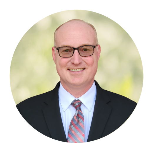 Portrait of a smiling man in a dark suit, glasses, and patterned tie against a soft green background