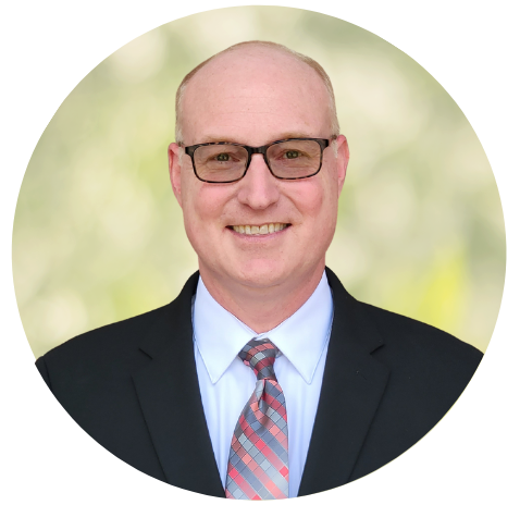Headshot of a smiling man in a black suit, light blue shirt, and plaid tie against a soft green background