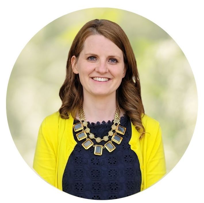 Smiling woman in a yellow cardigan and navy top, wearing a gold statement necklace, against a blurred green background