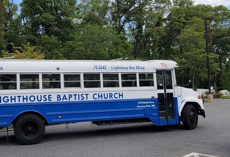 White and blue Lighthouse Baptist Church bus parked on a tree-lined street