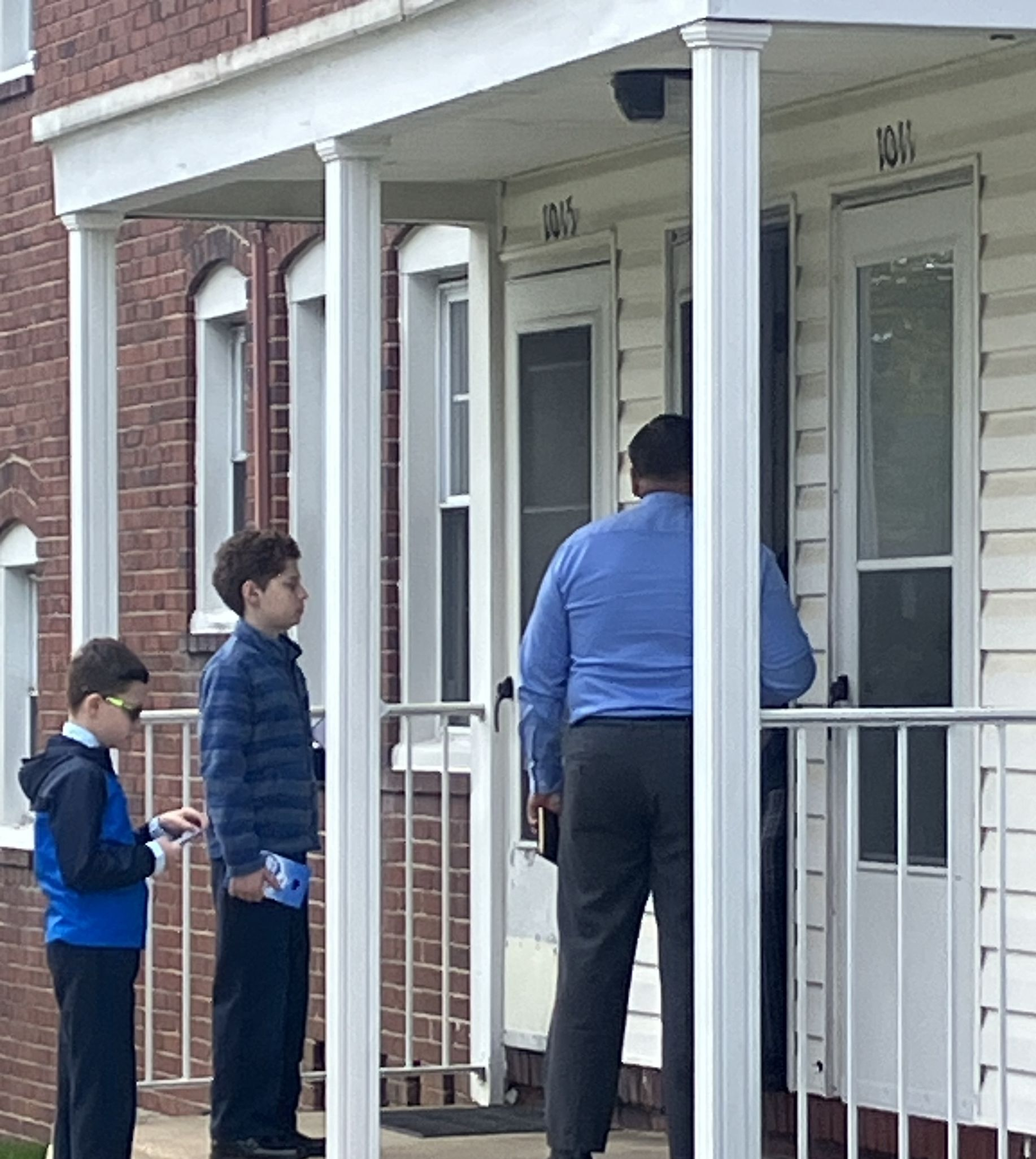 Three people on a porch at a house, one holding a package near the front door.