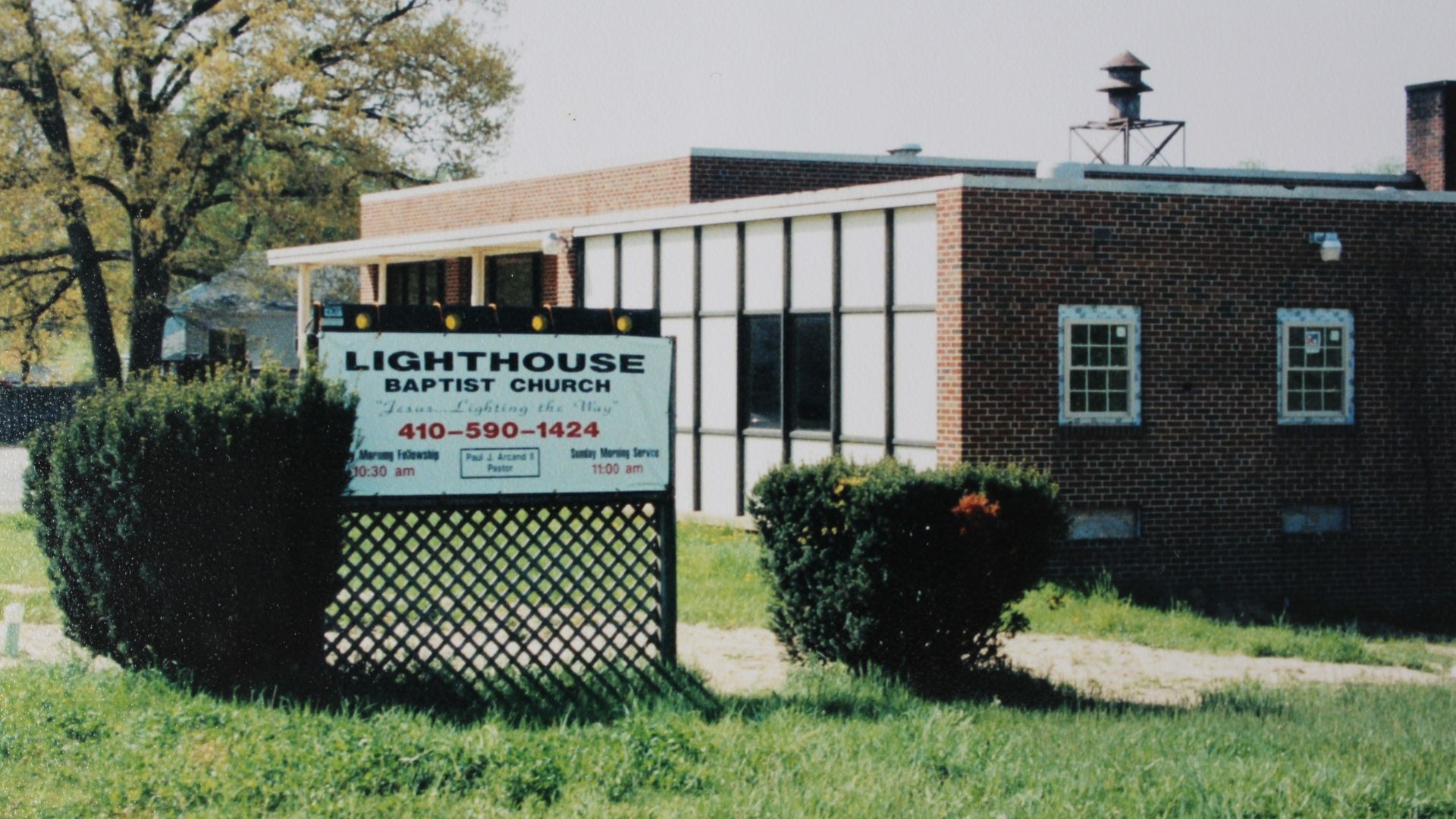 Lighthouse sign outside a brick building, with bushes and a tree in the foreground