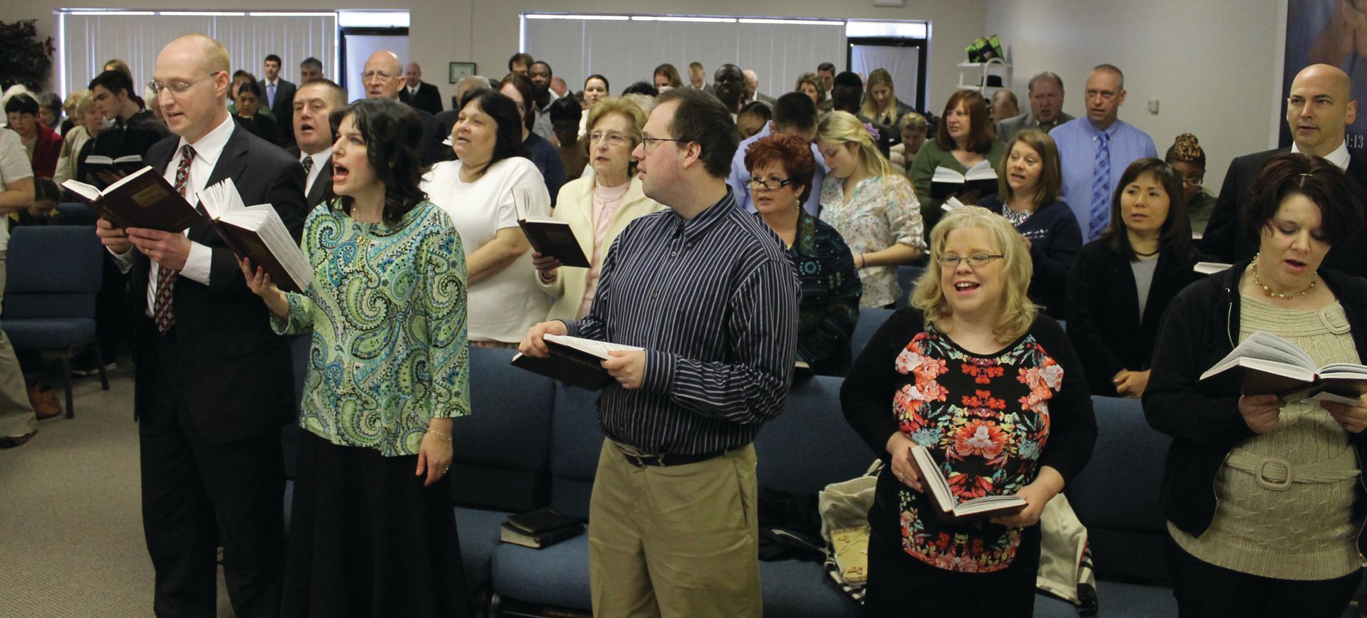 People standing and singing in a crowded indoor gathering, holding booklets and papers.