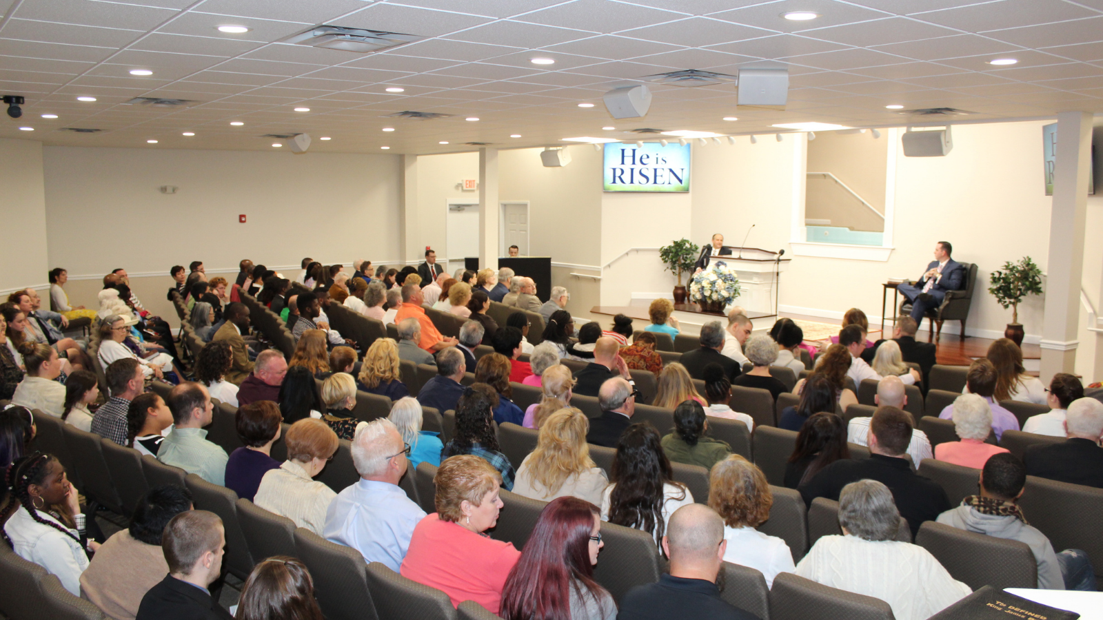 Large audience seated in a bright lecture hall, facing a speaker at the front near a screen.