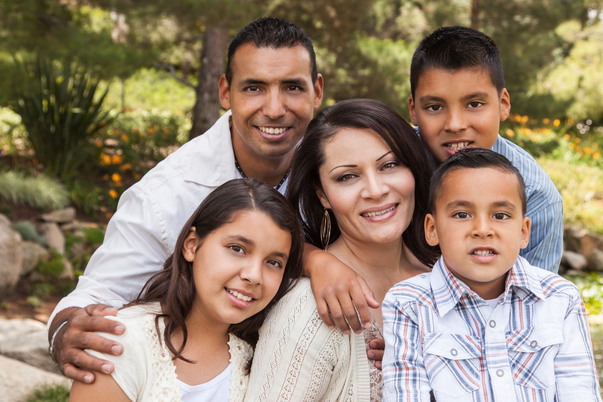Family portrait: Father with arm around mother, children smiling. Outdoors, greenery.