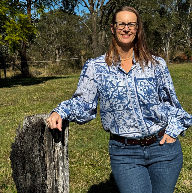 Woman in blue patterned shirt leaning on a weathered post in a sunny grassy field