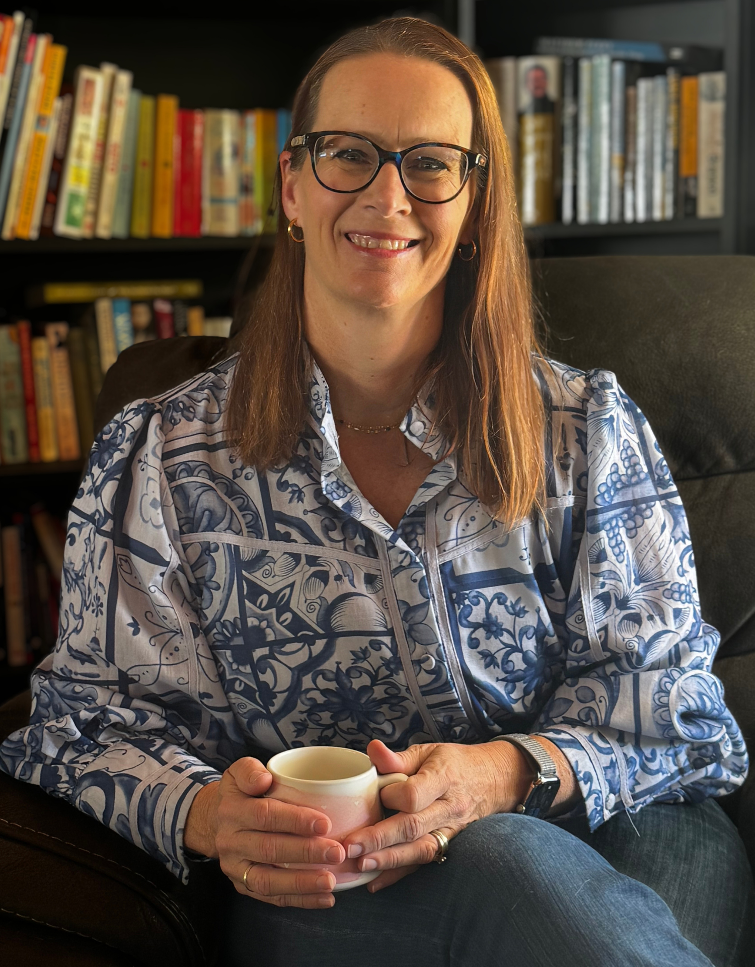 Woman seated in an armchair, smiling and holding a mug in front of a bookshelf.