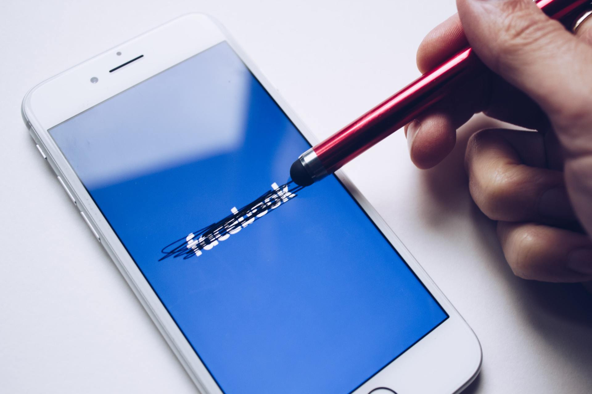 A person is holding a pen over a cell phone with the facebook logo on the screen.