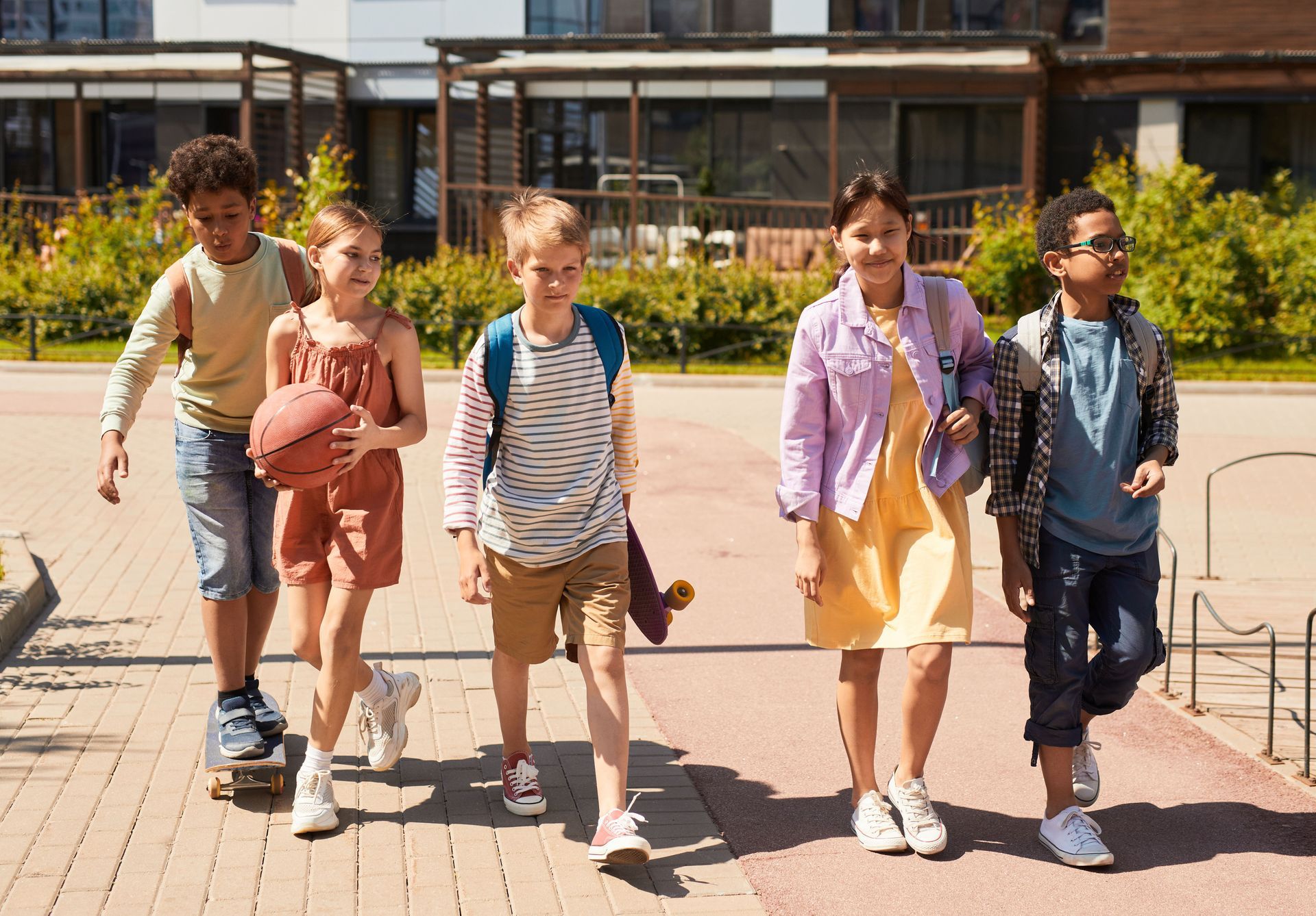 Five kids with backpacks walking together on a sunny sidewalk outside a school on the first day back