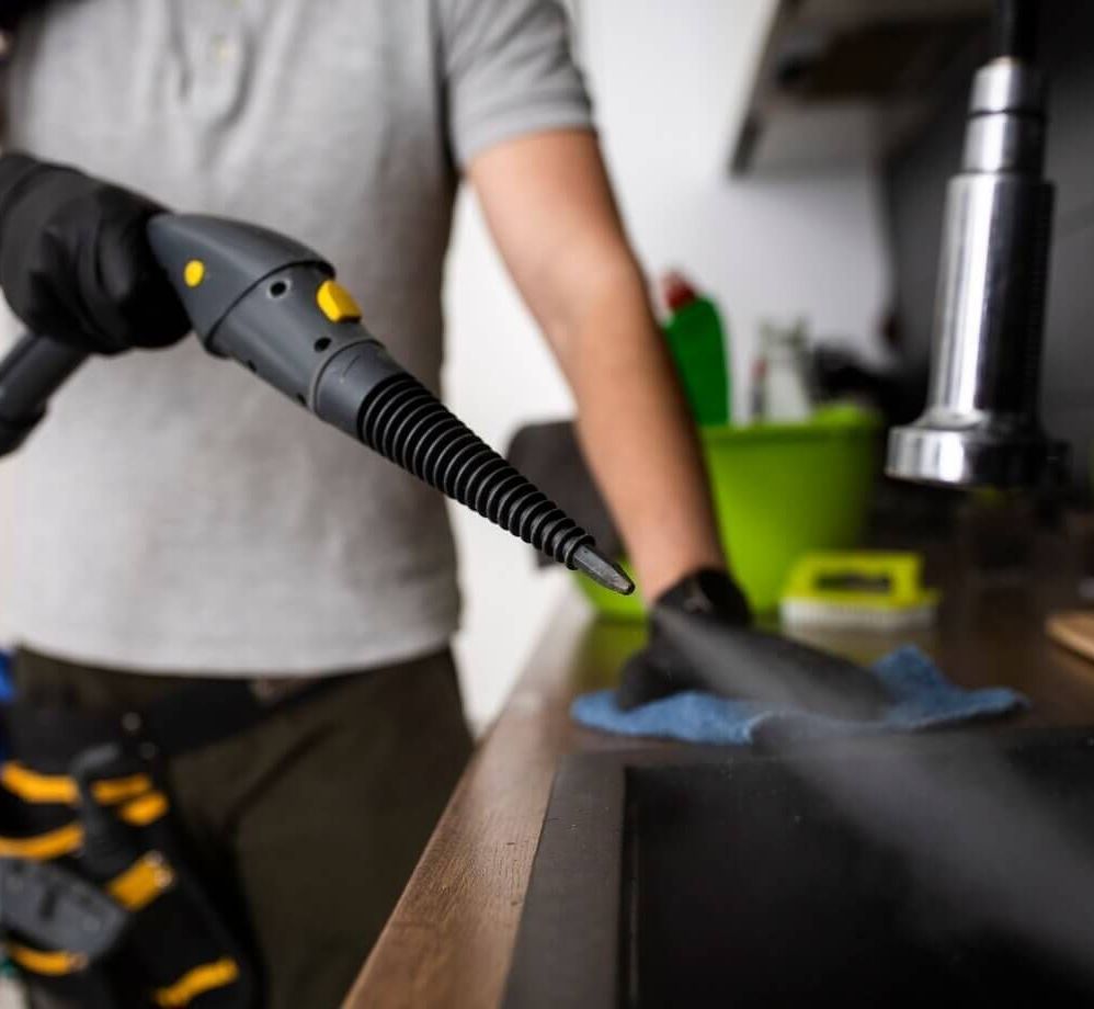 Un hombre está usando un limpiador a vapor para limpiar un fregadero en una cocina.