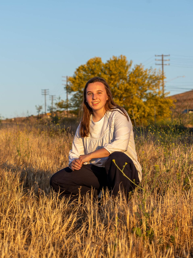 A person crouches in a field of golden dry grass during sunset, wearing a white long-sleeved shirt and dark pants.