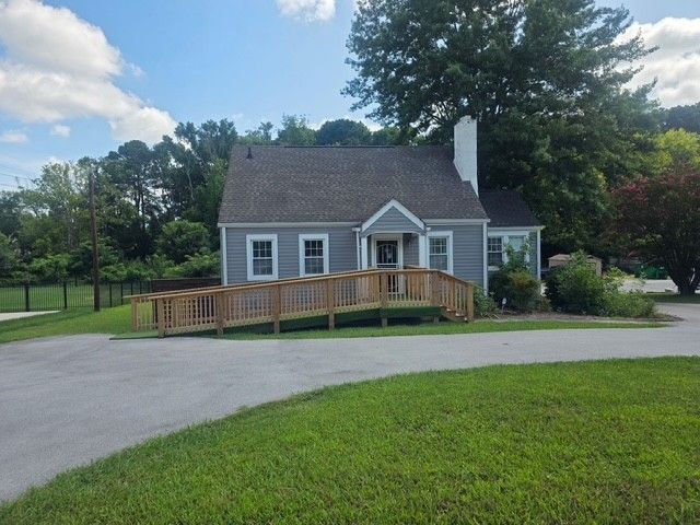 Small gray house with a wooden ramp, surrounded by trees and a curved driveway.