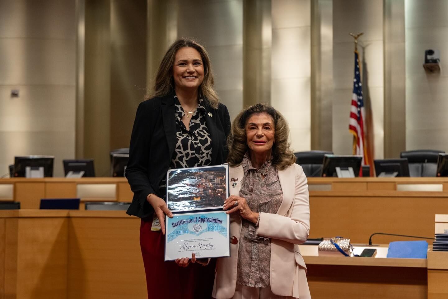 Two women pose in a council chamber holding a certificate and photo collage, with an American flag behind them.