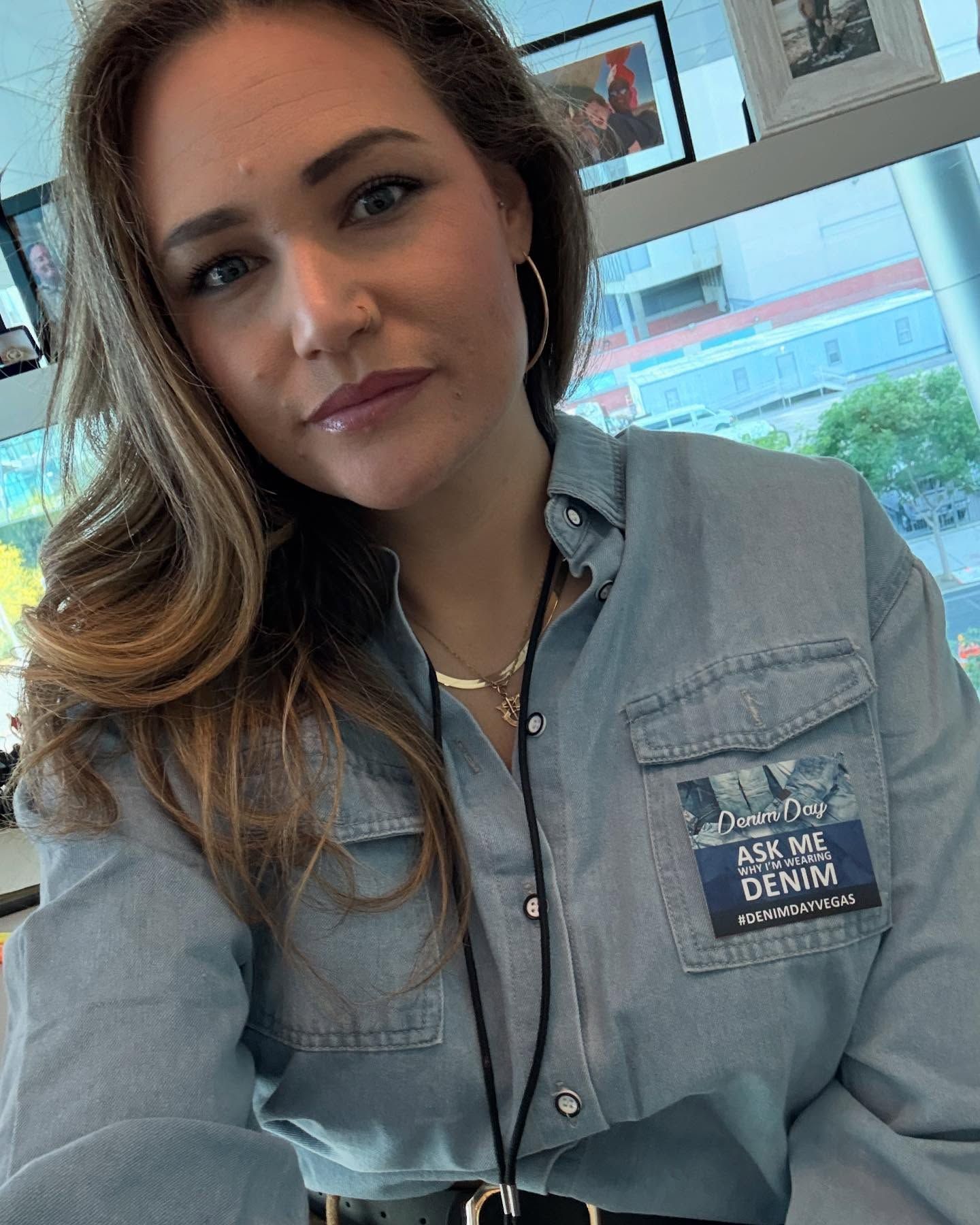 Woman in a denim shirt taking a selfie by a window in a bright indoor setting.