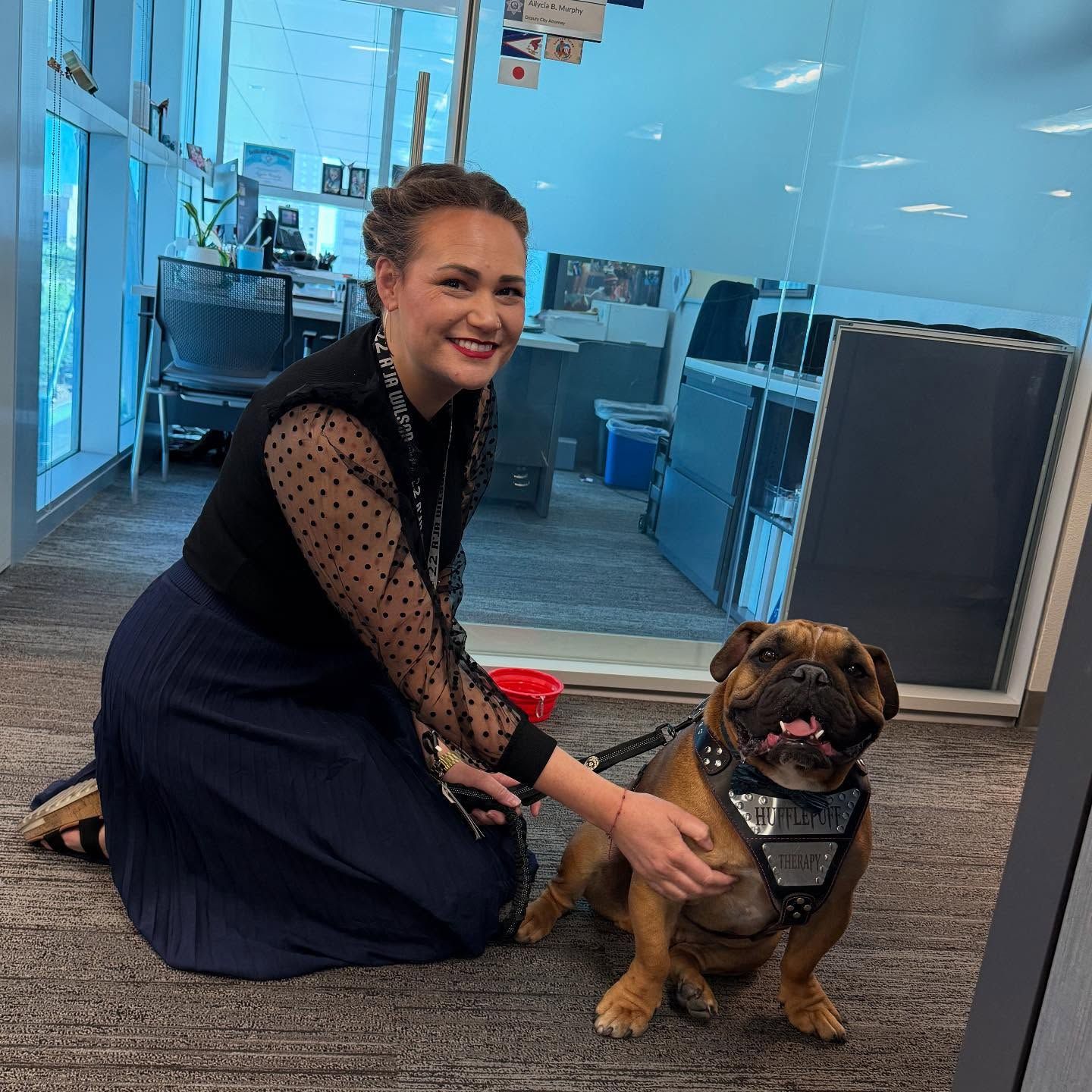 Woman kneeling beside a bulldog in an office, smiling as the dog sits on carpet.