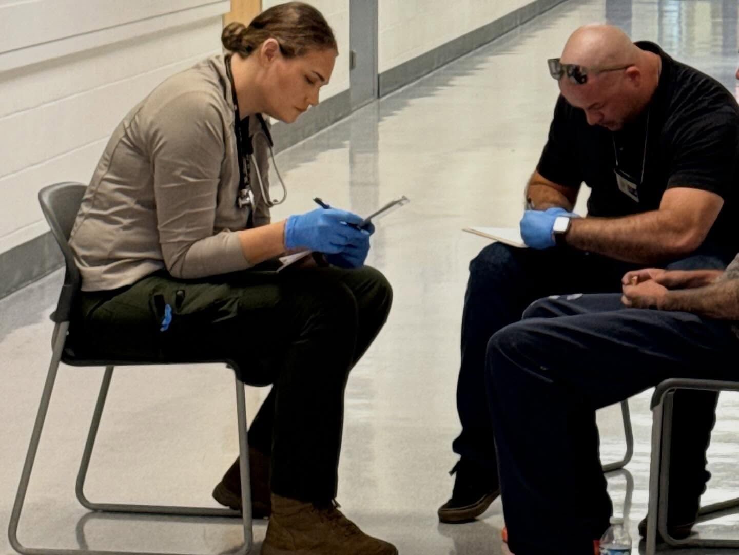 Two seated people in gloves examine a small item on a hallway floor.