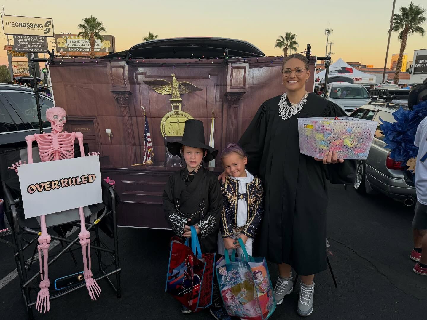 Costumed group posing by a decorated car at dusk, holding colorful treat bags and signs.