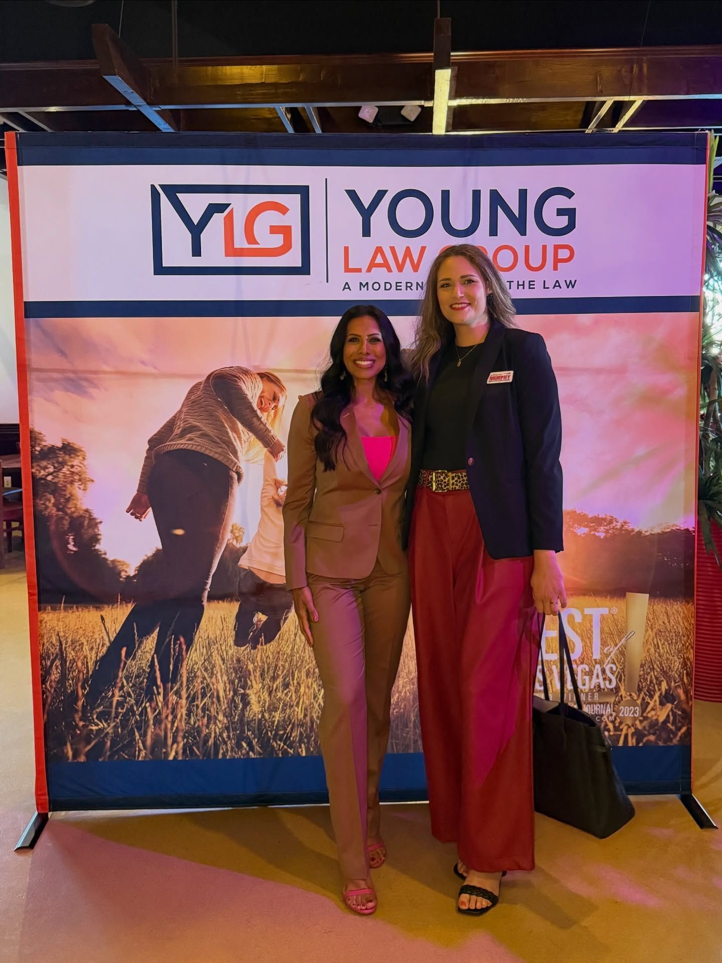 Two women posing at a Young Law Group event banner, smiling in front of a scenic backdrop.
