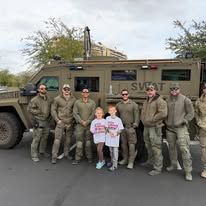 Group in military uniforms posing beside an armored vehicle, with two children holding signs in front