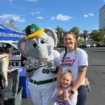 Mascot in white-and-green uniform posing with a woman and child at an outdoor event under a blue sky