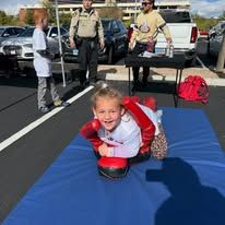 Child crawling on a blue mat in a parking lot, smiling, with adults and cars in the background