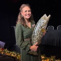 Smiling woman in a green dress holding a large fish trophy on a stage