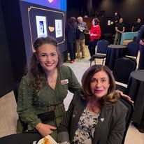 Two women smiling at a conference table in a dimly lit event space with people and posters in the background
