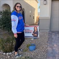 Person standing by a political yard sign in front of a house, wearing sunglasses and a blue jacket