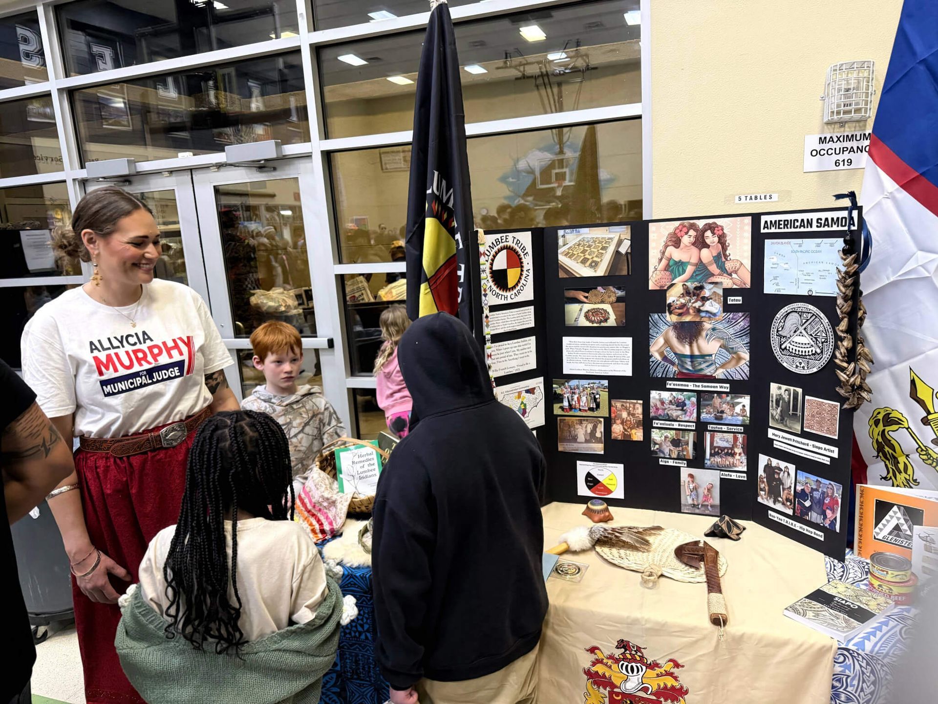 Student fair booth with poster display and people talking around a table in a gym-like room