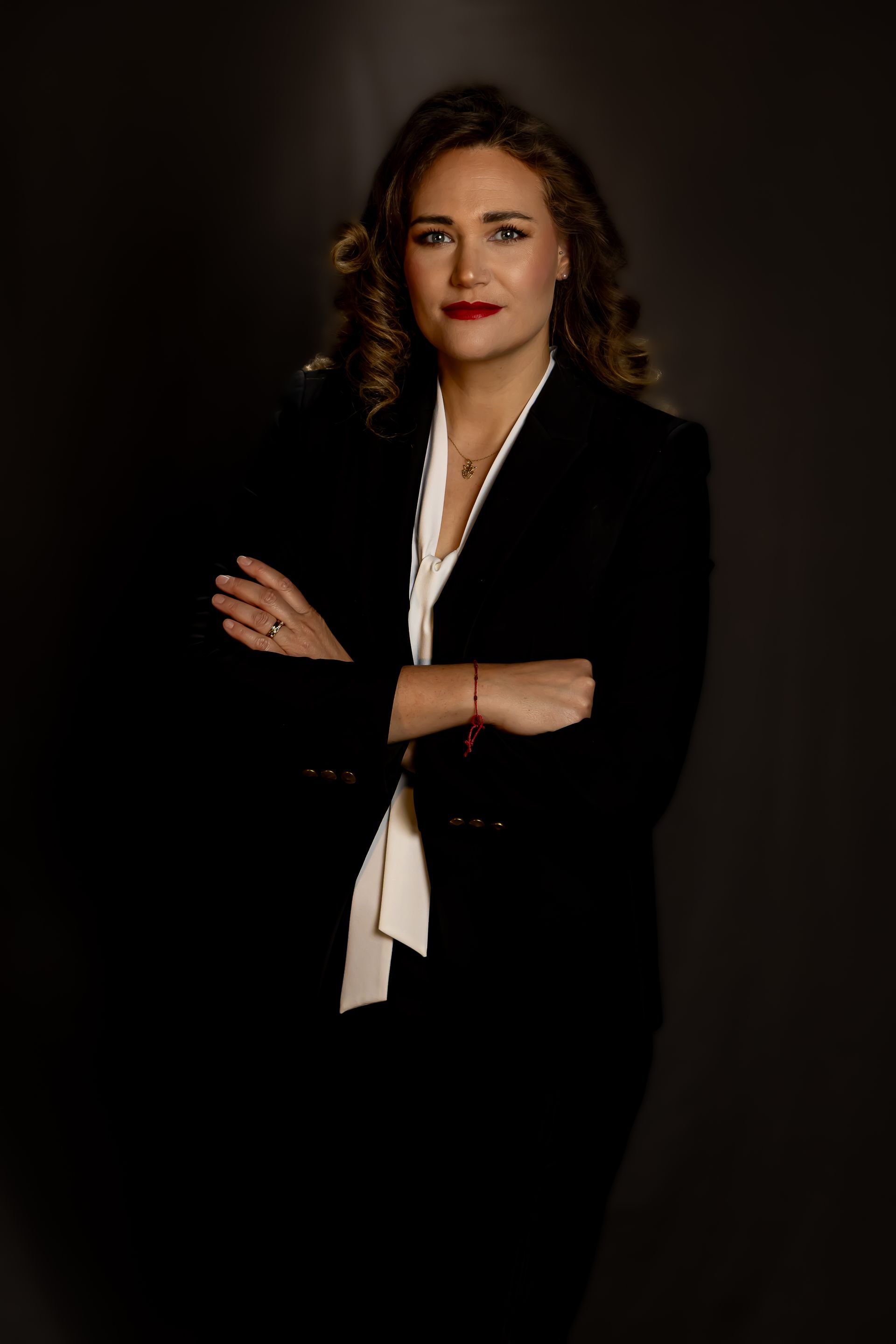 Woman in a black blazer with arms crossed, posing against a dark studio background.