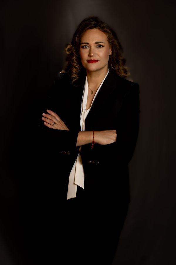 Woman in a black blazer with arms crossed, posing against a dark studio background.