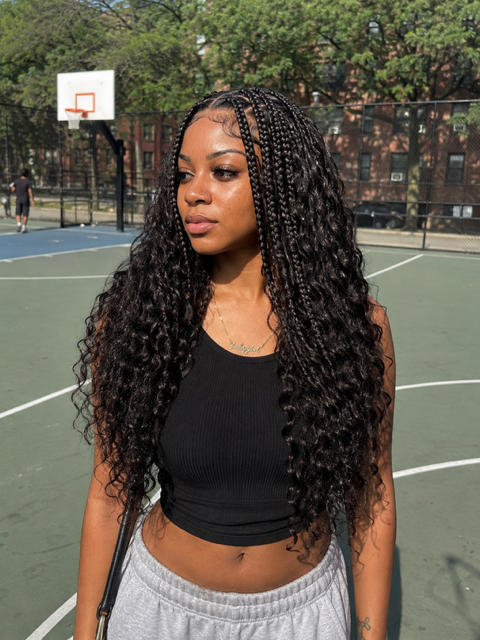 Woman standing on an outdoor basketball court, wearing a black top and gray pants, with long braided hair.