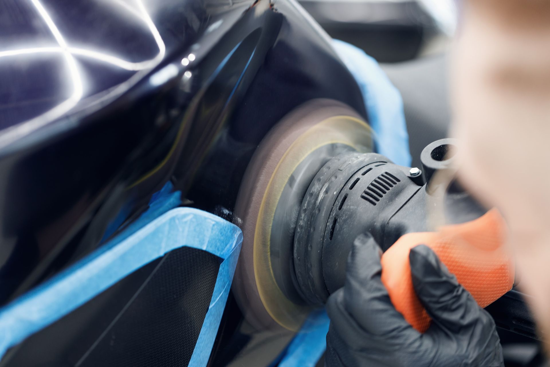 Gloved hand polishing a black car with a rotary buffer and orange pad