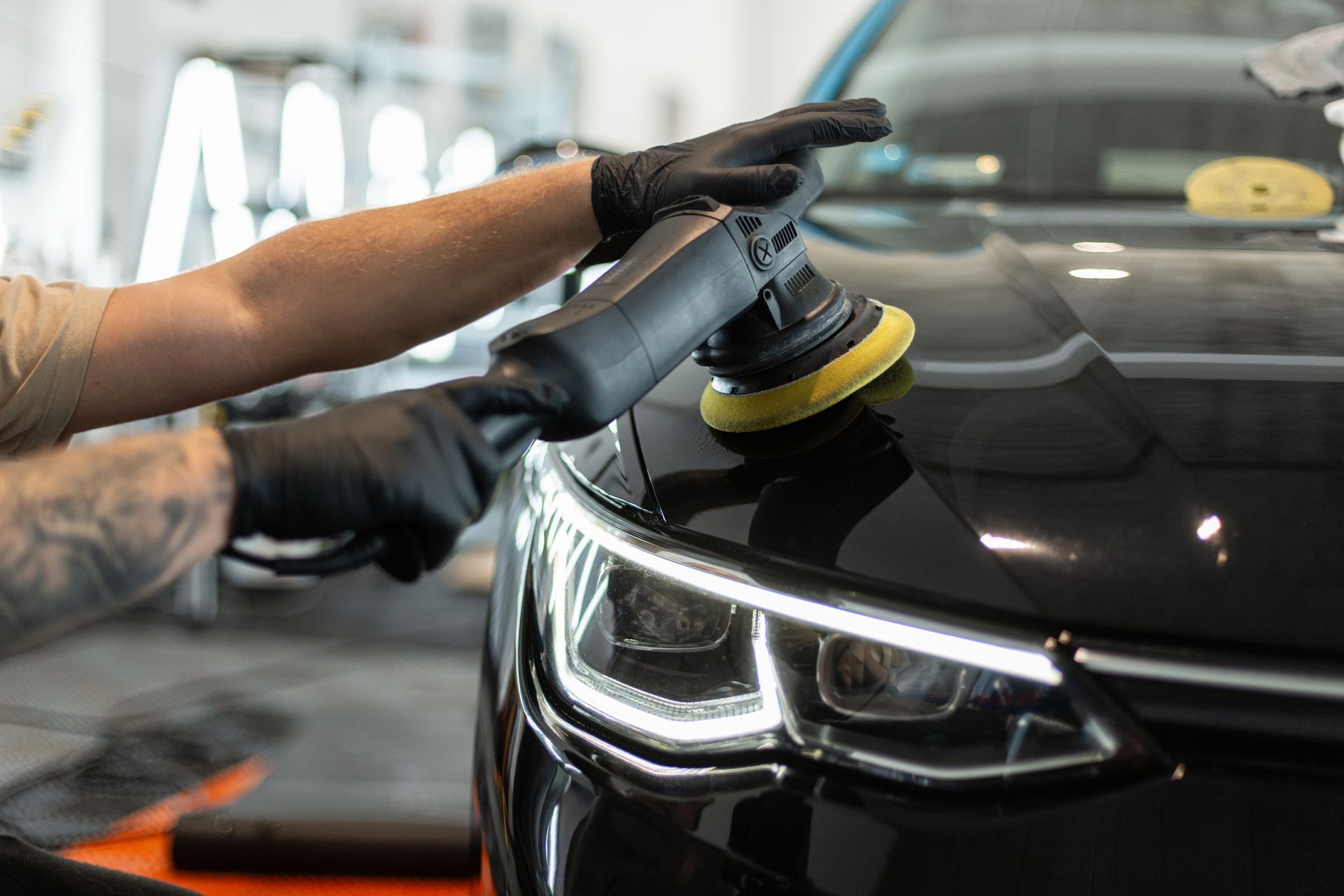 Detailer polishing a black car hood with a buffer in a garage