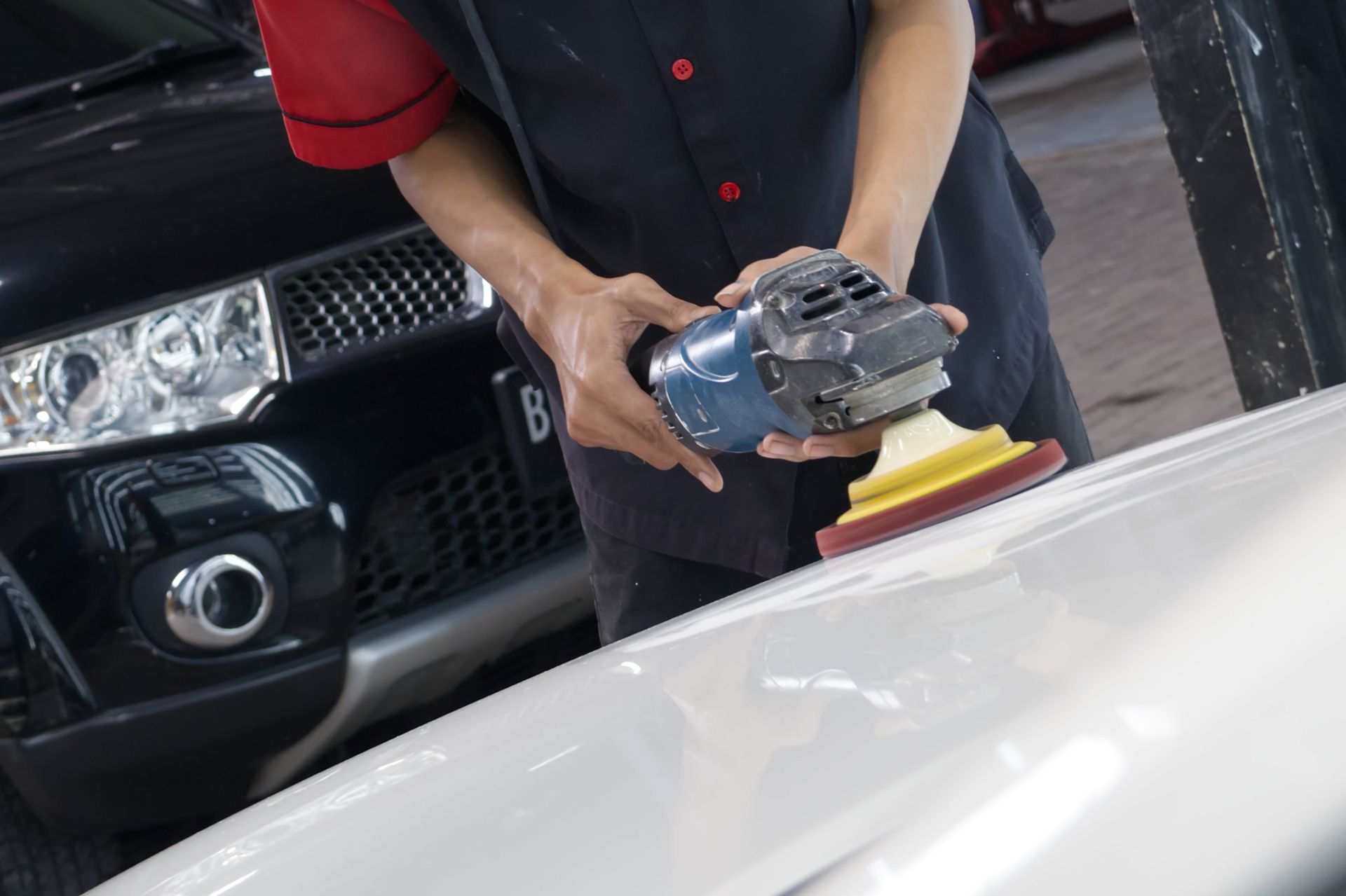 Person using a power sander on a white car hood beside a black vehicle