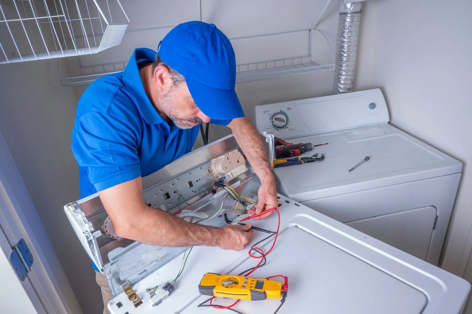 A repairman in a blue uniform working on a disassembled white dryer, using tools in a utility room.